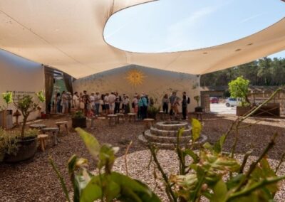 Groupe de personnes arrivant dans une ferme à Ibiza, sous une toile tendue, entourés de bancs en bois, de plantes en pot et d’un mur décoré avec un soleil jaune et des motifs floraux.
